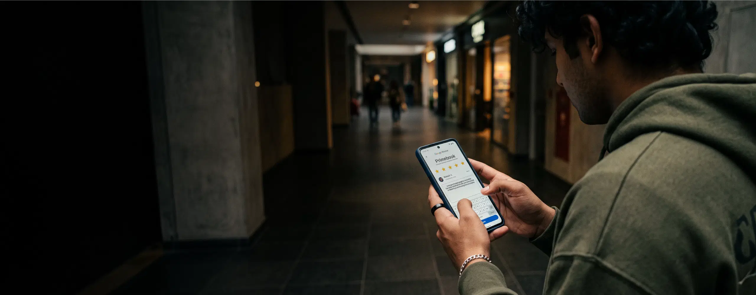 Man in a hoodie reading a five-star Primebook review on his phone in a dim corridor