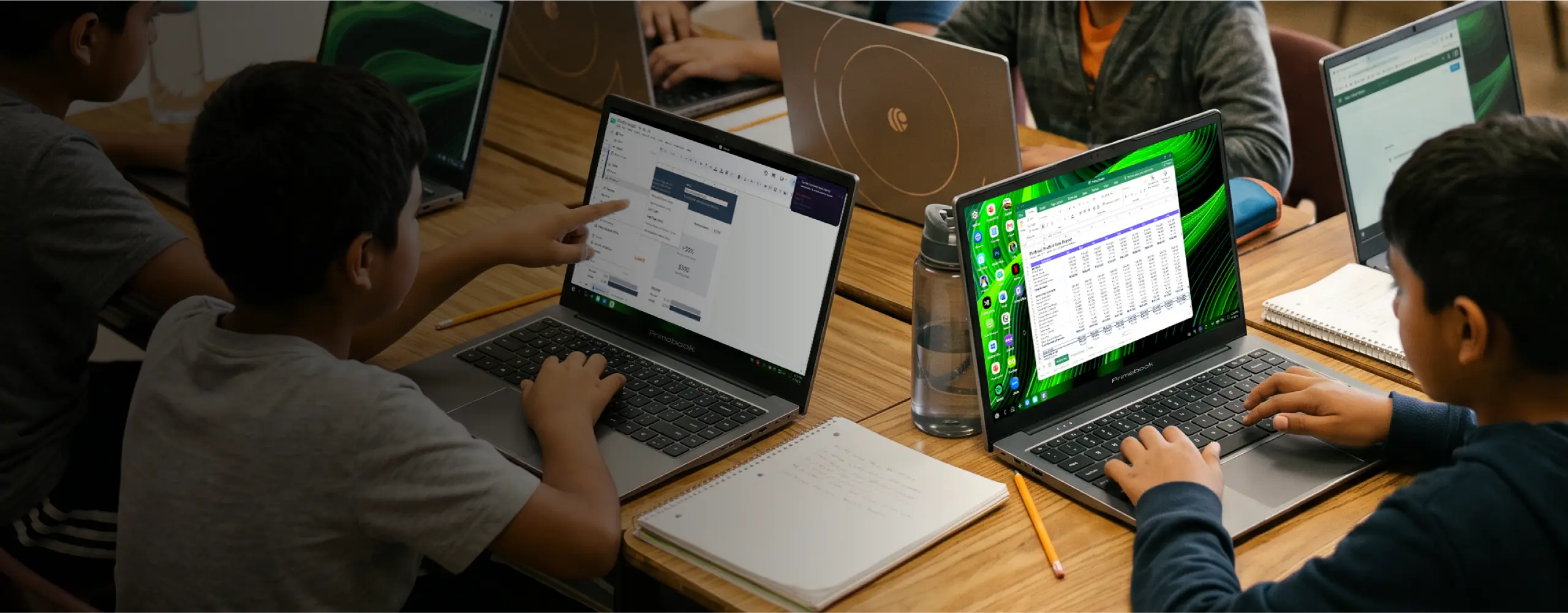 Children working on Primebook laptops around a classroom table with notebooks and pencils